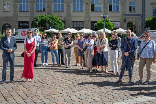 Grupa ludzi stojąca na placu, w różnorodnych strojach, w słoneczny dzień.