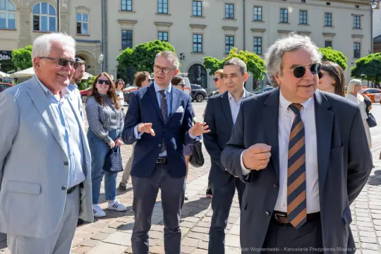 Grupa mężczyzn rozmawia na placu, w tle budynki i przechodnie.