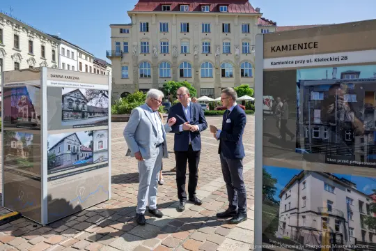 Trzech mężczyzn rozmawia przy wystawie zdjęć kamienic w mieście.
