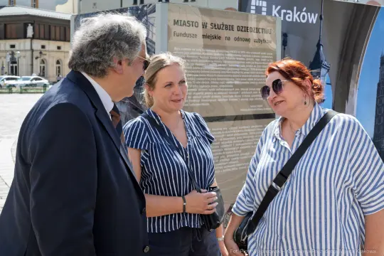 Trzy osoby rozmawiają na tle tablicy informacyjnej o funduszach miejskich w Krakowie.