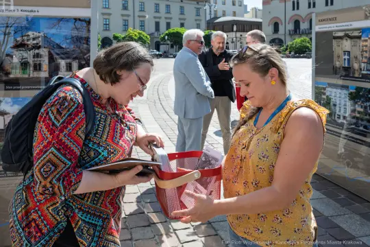 Dwie kobiety rozmawiają i przeglądają materiały w przestrzeni publicznej. W tle inni ludzie.
