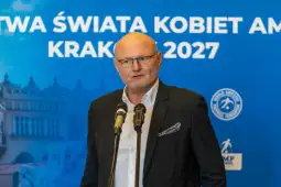 Mężczyzna przemawia do mikrofonów na tle niebieskiego banera promującego wydarzenie w Krakowie.