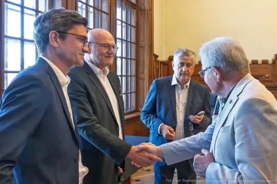 Spotkanie czterech mężczyzn, uściski dłoni i rozmowy w sali z dużymi oknami.
