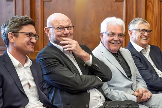 Czterech mężczyzn w garniturach siedzących obok siebie, uśmiechających się.