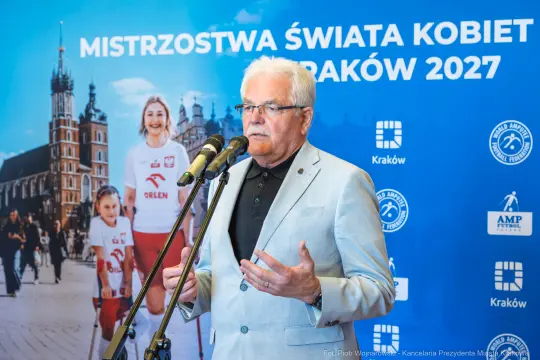 Mężczyzna w jasnym garniturze przemawia na tle plakatu Mistrzostw Świata Kobiet w Krakowie 2027.