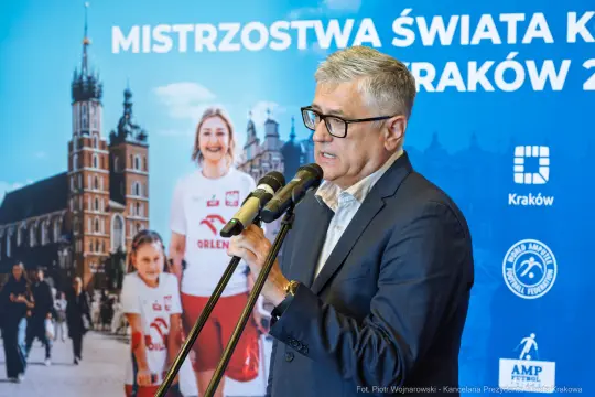 Mężczyzna przemawia przy mikrofonach, w tle plakat Mistrzostw Świata w Krakowie.
