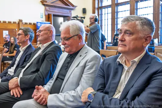 Czterech mężczyzn siedzących w sali, skupionych na wydarzeniu.