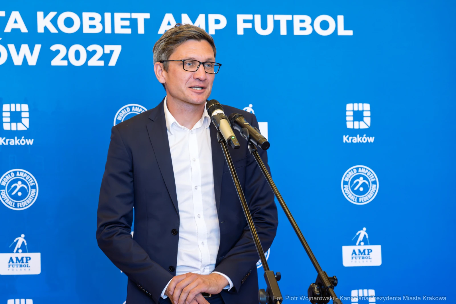Mężczyzna w garniturze przemawia do mikrofonu na tle niebieskiego tła z logo AMP Futbol.  Autor: Piotr Wojnarowski