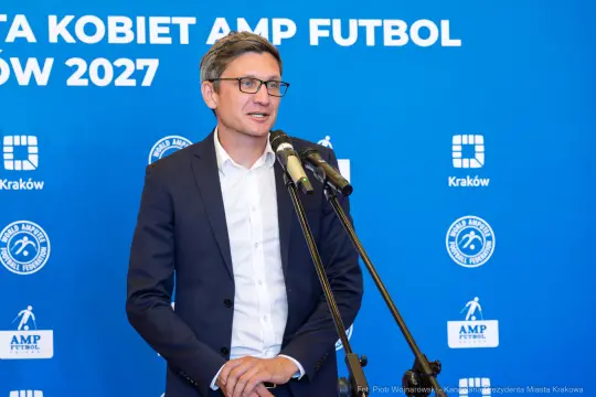 Mężczyzna w garniturze przemawia do mikrofonu na tle niebieskiego tła z logo AMP Futbol.