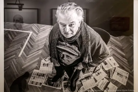 Mężczyzna w swetrze z nadrukiem, stojący na gazetach w artystycznym wnętrzu.