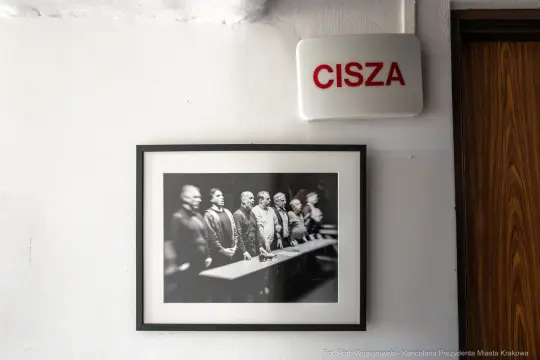 Czarnobiałe zdjęcie grupy ludzi stojących przy stole, obok napis "CISZA".