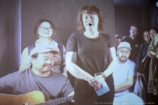 Kobieta śpiewająca, mężczyzna grający na gitarze, grupa osób w tle.