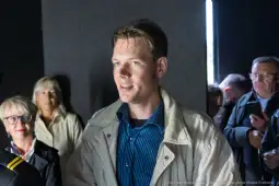 Mężczyzna w jasnej kurtce i niebieskiej koszuli, otoczony przez grupę ludzi.