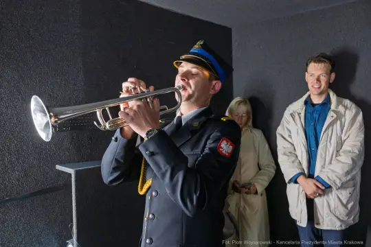Mężczyzna w mundurze gra na trąbce, obok dwie osoby słuchają.