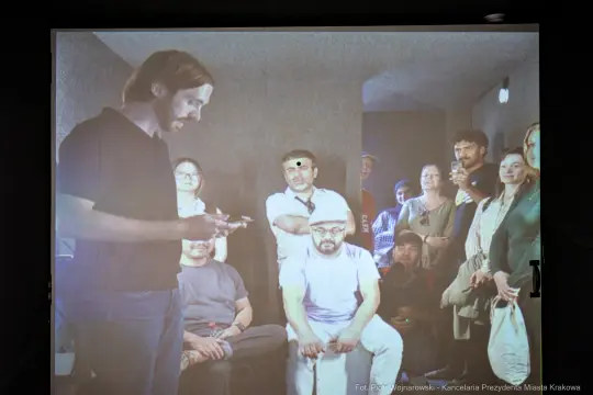 Mężczyzna prezentujący coś grupie ludzi w zamkniętej przestrzeni.