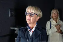 Kobieta w okularach z blond włosami, w eleganckim płaszczu, słucha uważnie. W tle druga kobieta.