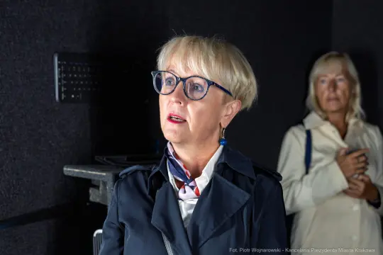 Kobieta w okularach z blond włosami, w eleganckim płaszczu, słucha uważnie. W tle druga kobieta.