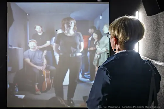 Kobieta obserwująca grupę ludzi w pomieszczeniu, w tym mężczyznę z gitarą.