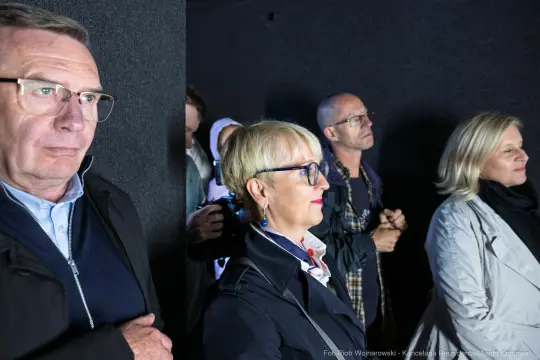 Grupa osób w zamkniętej przestrzeni, skupionych na czymś przed sobą.
