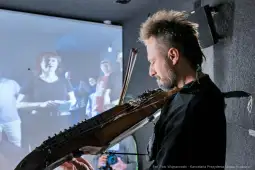 Muzyk grający na instrumentach strunowych, w tle widoczna grupa ludzi.