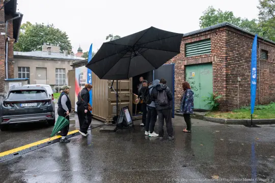 Ludzie stojący pod parasolem przy kontenerze w deszczowy dzień.