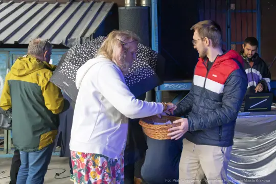 Osoba z parasolem odbiera koszyk od mężczyzny w kurtce, w tle inni uczestnicy wydarzenia.