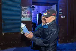 Starszy mężczyzna trzyma butelkę, obok niego kobieta w tle. Wnętrze z niebieskim oświetleniem.