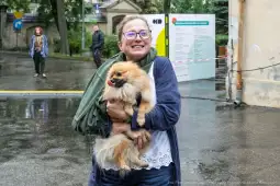 Kobieta trzyma w ramionach psa rasy pomeranian, uśmiechnięta, w deszczowej scenerii.