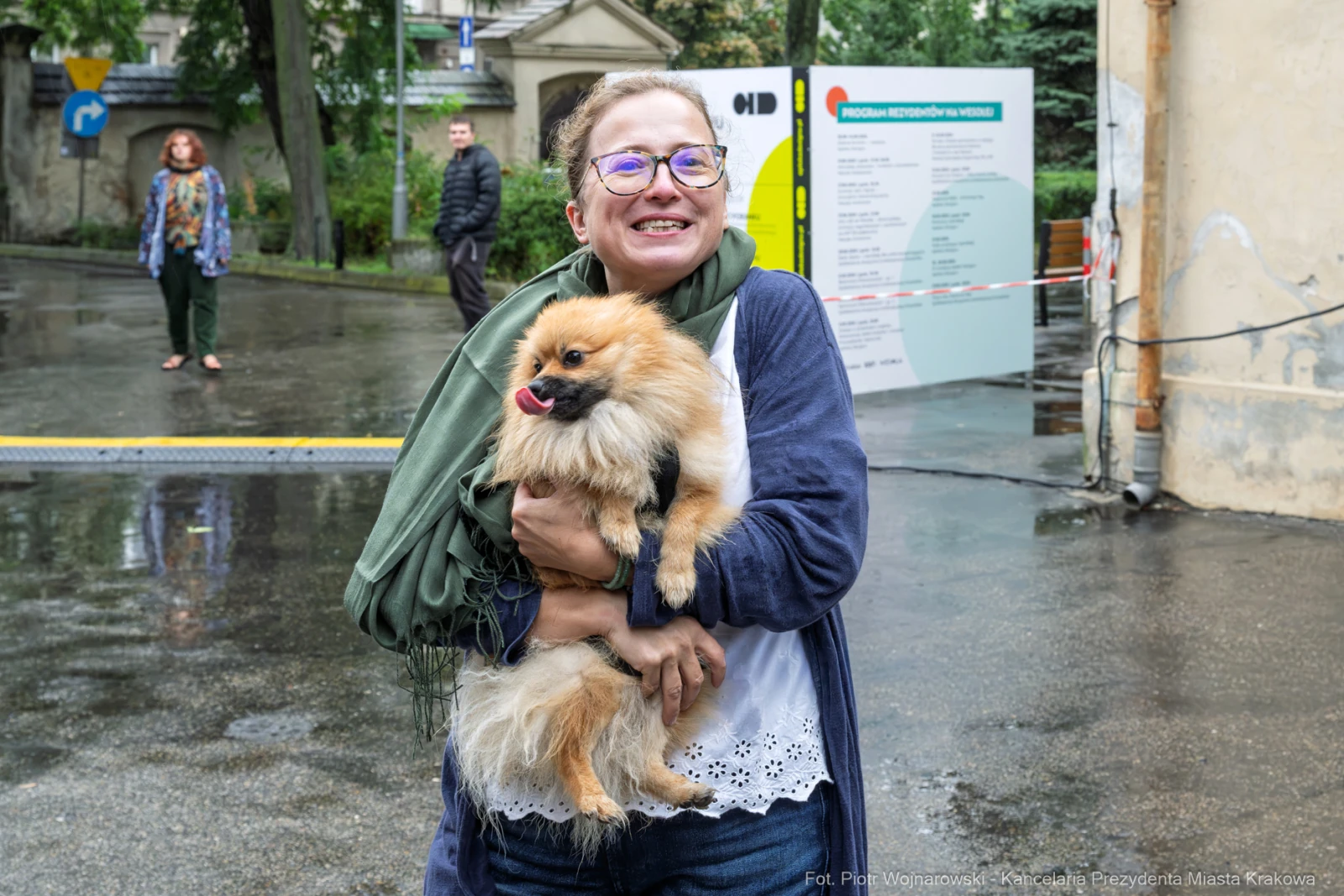 Kobieta trzyma w ramionach psa rasy pomeranian, uśmiechnięta, w deszczowej scenerii.  Autor: Piotr Wojnarowski