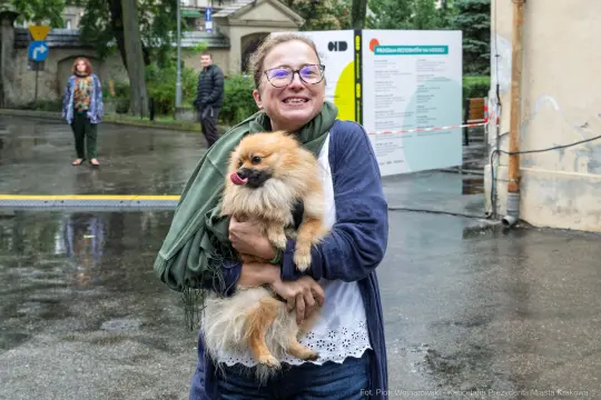 Kobieta trzyma w ramionach psa rasy pomeranian, uśmiechnięta, w deszczowej scenerii.