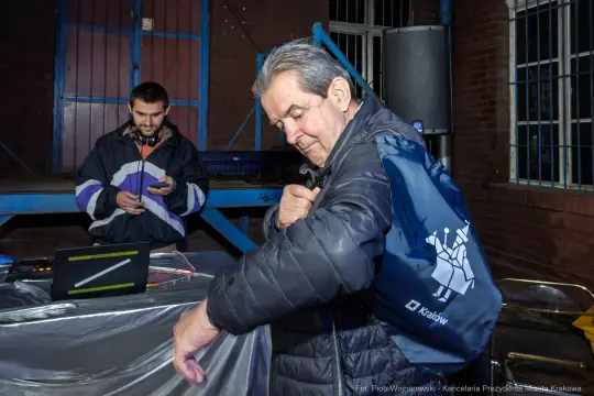 Mężczyzna z torbą na ramieniu, w tle inny mężczyzna przy laptopie.