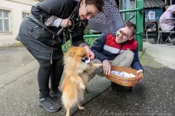 Kobieta z parasolką podaje kartkę mężczyźnie, obok nich stoi mały pies.