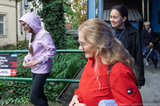 Grupa osób spacerujących w miejskim otoczeniu, jedna z nich w czerwonej kurtce.