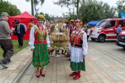Dwie kobiety w tradycyjnych strojach ludowych z kwiatowymi wiankami, niosące wieńce dożynkowe.