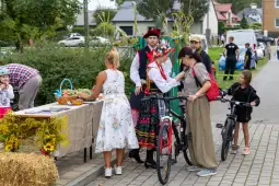 Kobiety w tradycyjnych strojach ludowych, stoiska z jedzeniem, rowerzyści i dzieci bawiące się na festynie.
