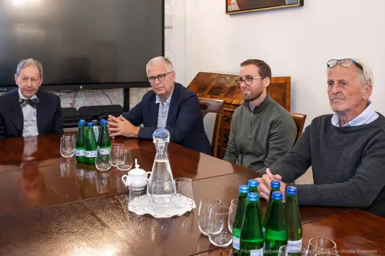 Czterech mężczyzn siedzących przy stole, obok butelki wody i szklanek.