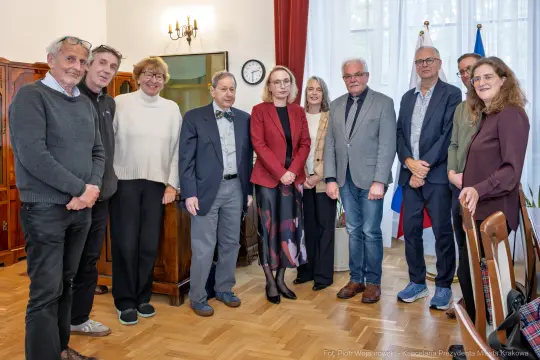Grupa osób w formalnych strojach w sali z flagami i zegarem.