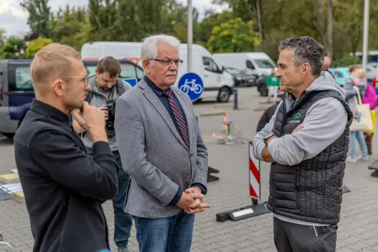 Trzech mężczyzn rozmawia na tle pojazdów i znaków drogowych.