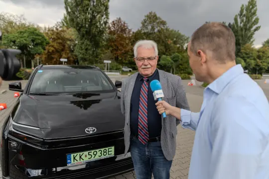 Mężczyzna w szarym garniturze i krawacie stoi obok czarnego samochodu Toyota, rozmawia z dziennikarzem.