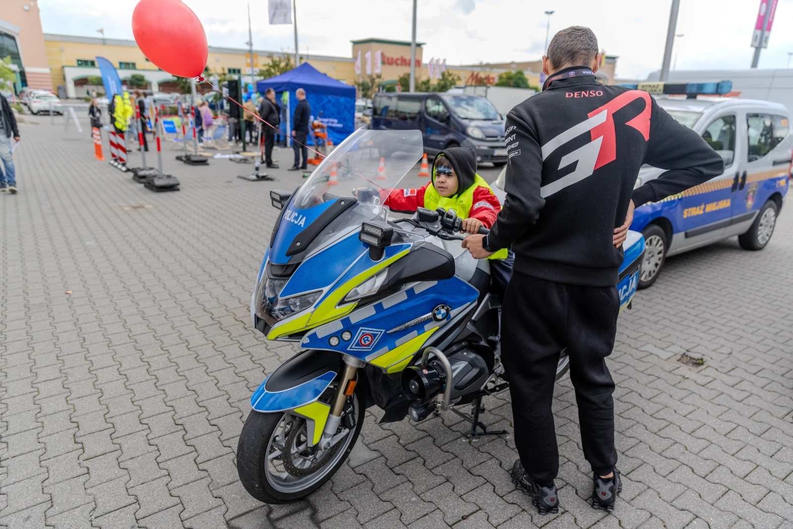 Dziecko siedzące na policyjnym motocyklu, obok dorosły w czarnej bluzie. W tle balon i stoiska.  Autor: Bogusław Świerzowski