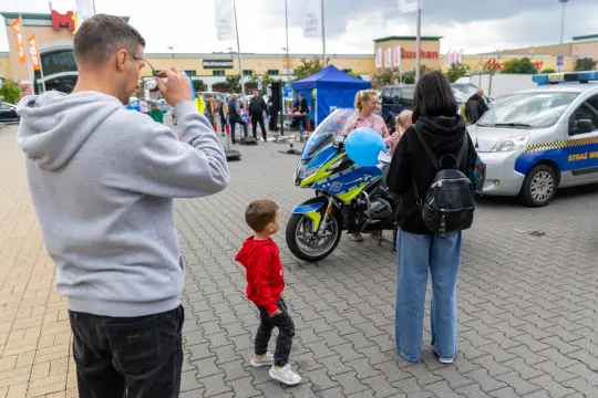 Dziecko w czerwonym kapturku obok motocykla policyjnego, dorośli rozmawiają z przedstawicielami służb.