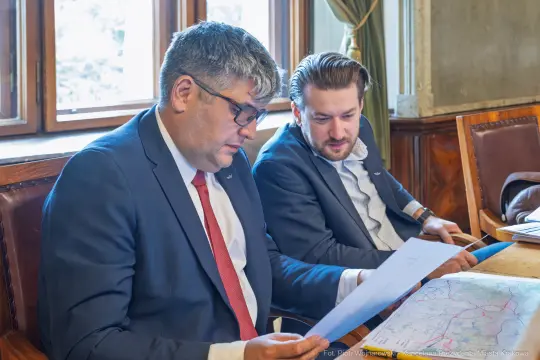 Dwóch mężczyzn w garniturach przegląda dokumenty przy stole w biurze.