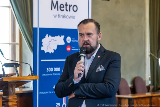 Mężczyzna z mikrofonem przemawia na tle plakatu dotyczącego metra w Krakowie.