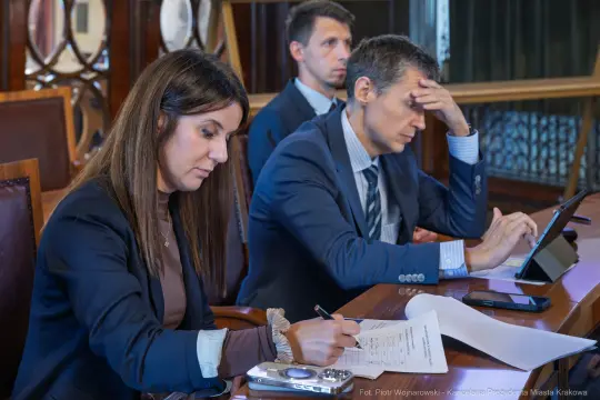 Trzy osoby w garniturach podczas spotkania, jedna notuje, dwie skupione na zadaniach.