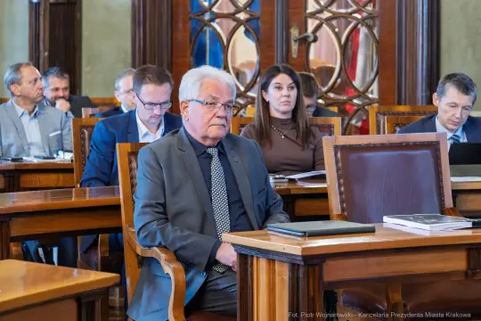 Mężczyzna w garniturze siedzi w sali obrad, w tle inni uczestnicy spotkania.