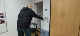 Mężczyzna z chodzikiem wyciera powierzchnię mebla w jasnym pomieszczeniu.