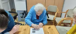 Starsza kobieta w niebieskim swetrze wypełnia formularz przy stole, obok dwie inne osoby.