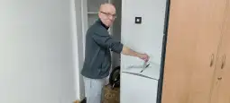 Mężczyzna w szarej bluzie stoi obok lodówki, trzymając kartkę w ręku.
