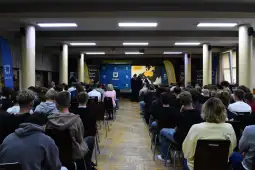 Wielka sala z tłumem ludzi siedzących na krzesłach, na scenie prezentacja lub wykład.