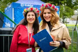 Dwie kobiety w kolorowych wiankach, uśmiechnięte, trzymają teczkę z logo Krakowa.